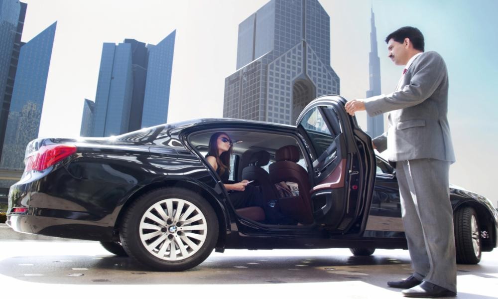 A chauffeur in a gray suit holds the door open for a woman sitting inside a luxury black car, with modern skyscrapers in the background